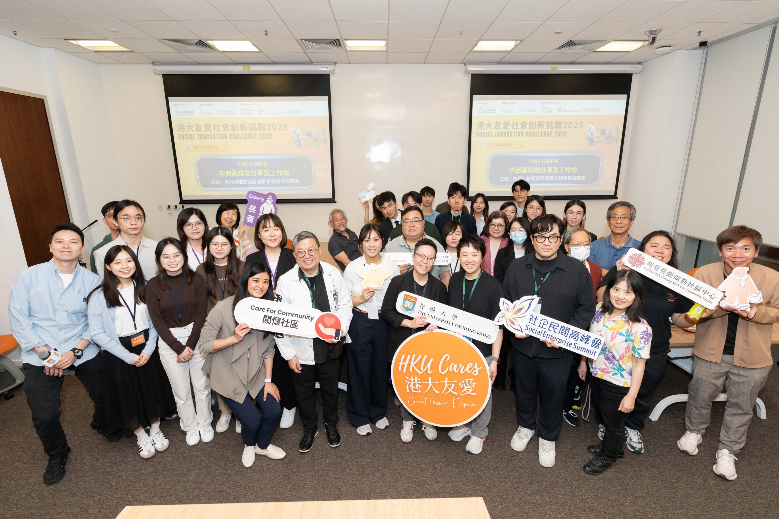Group photo of participants and organizers at the HKU Social Innovation Challenge 2025, holding banners and signs promoting community care, social enterprise, and innovative collaborations, with event details displayed on presentation screens in the background.