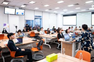 A speaker addresses an engaged audience in a modern classroom setting at HKU, featuring collaborative group tables, orange chairs, and presentation screens displaying event content