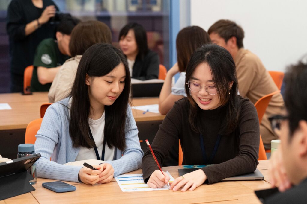 Two students collaborate and share ideas while writing on a worksheet during a group activity at the HKU Social Innovation Challenge 2025, surrounded by other engaged participants.