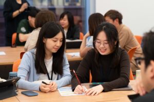 Two students collaborate and share ideas while writing on a worksheet during a group activity at the HKU Social Innovation Challenge 2025, surrounded by other engaged participants.