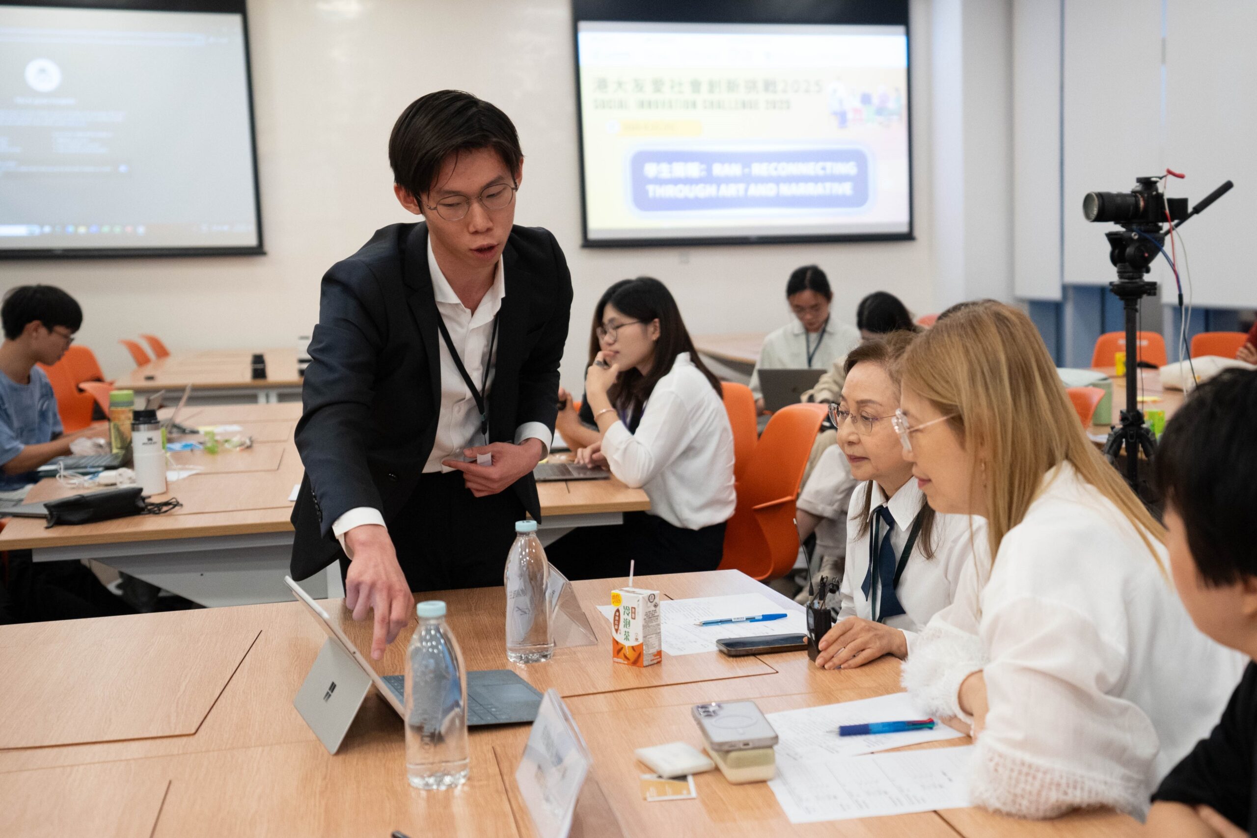 A participant in a suit explains a concept to a group of attentive attendees seated at a table during the HKU Social Innovation Challenge 2025, with a presentation screen and collaborative workspace in the background.